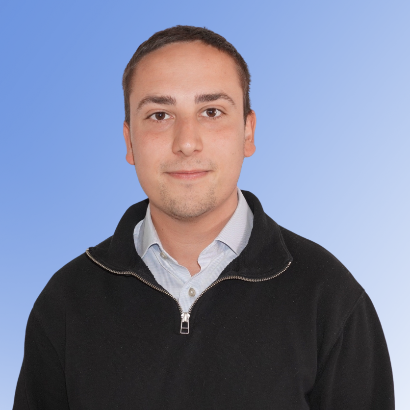 Yani Carter, Senior Consultant at ACE Partners, wearing a black zip sweater and collared shirt on a blue background.
